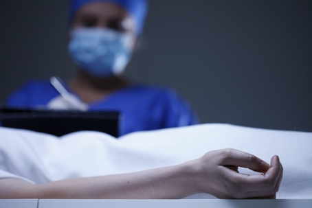 A patient’s hand rests motionless on a hospital bed while a masked healthcare worker stands in the background, suggesting a hospital death or serious medical outcome.